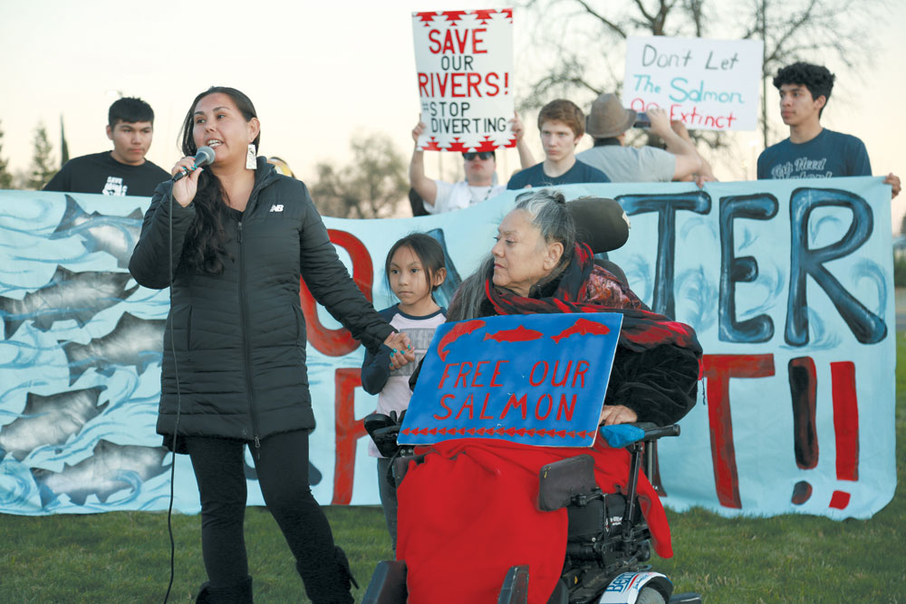 A woman standing at mic, with child beside her, and an elder woman in a wheelchair holding a painted sign reading "Free our Salmon" Students hold a banner behind them reading "No More Water for Profit!"