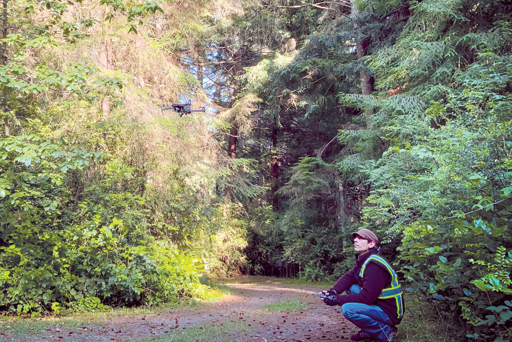 man in woods squatting down looking up at flying drone in the foreground.