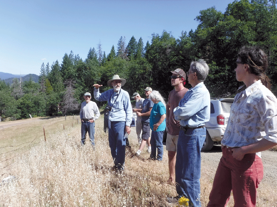 eight people standing in field with forest in background, one man in sun hat speaking.