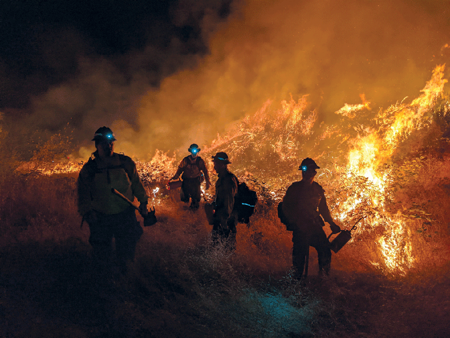 four figures in prescribed burn gear wearing helmets with headlights walk beside a large nighttime prescribed burn.
