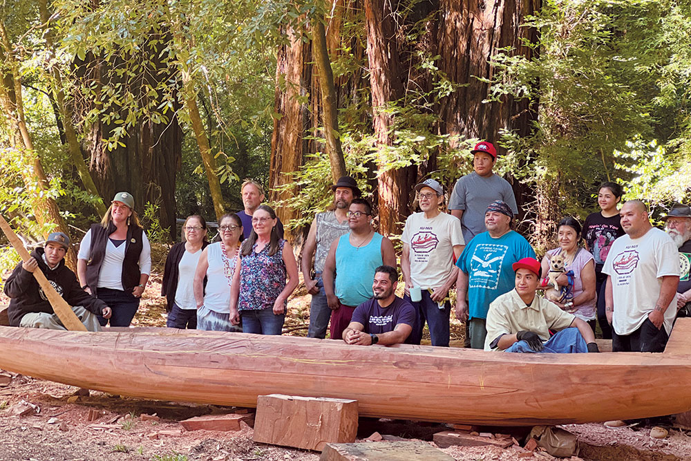 Ernest Lincoln, on the far left, holds a canoe paddle and sits in the canoe that he and other Wailaki are building as a renewal of an ancient cultural tradition. To his right is Erin Gates, the Deputy District Superintendent for California State Park’s North Coast Redwood District, along with other participants and supporters of the redwood canoe project. 